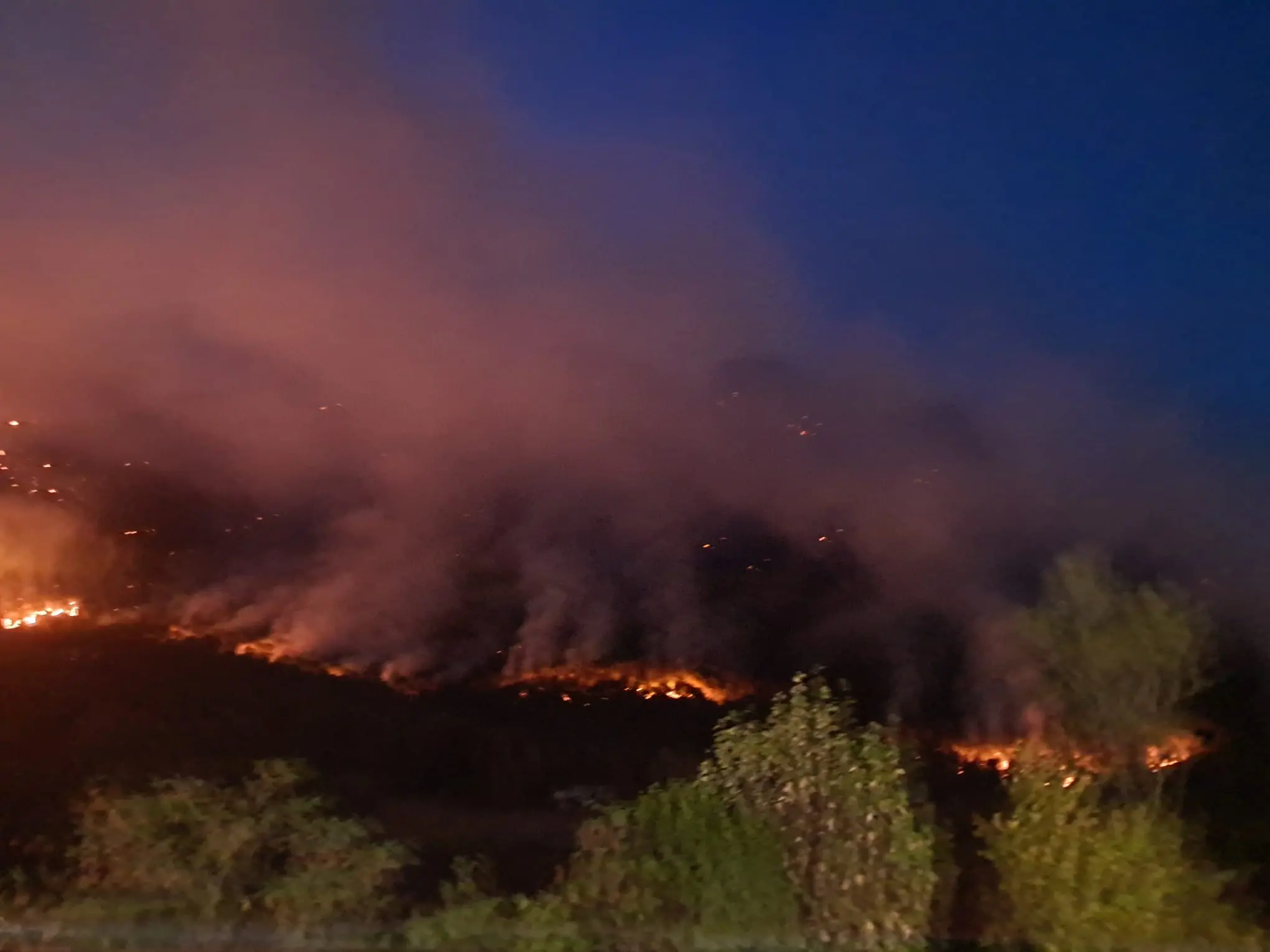 Le colonne di fumo degli incendi che stanno bruciando in Carnia sono visibili a chilometri di distanza e in alcune zone l’aria è diventata difficile da respirare. Una coltre grigia avvolge vallate e montagne, oscurando il cielo e cambiando il volto del paesaggio. Nella notte appena trascorsa, nel buio erano le fiamme sui versanti a illuminare la montagna, mentre con il giorno il fumo è tornato a dominare lo scenario.
