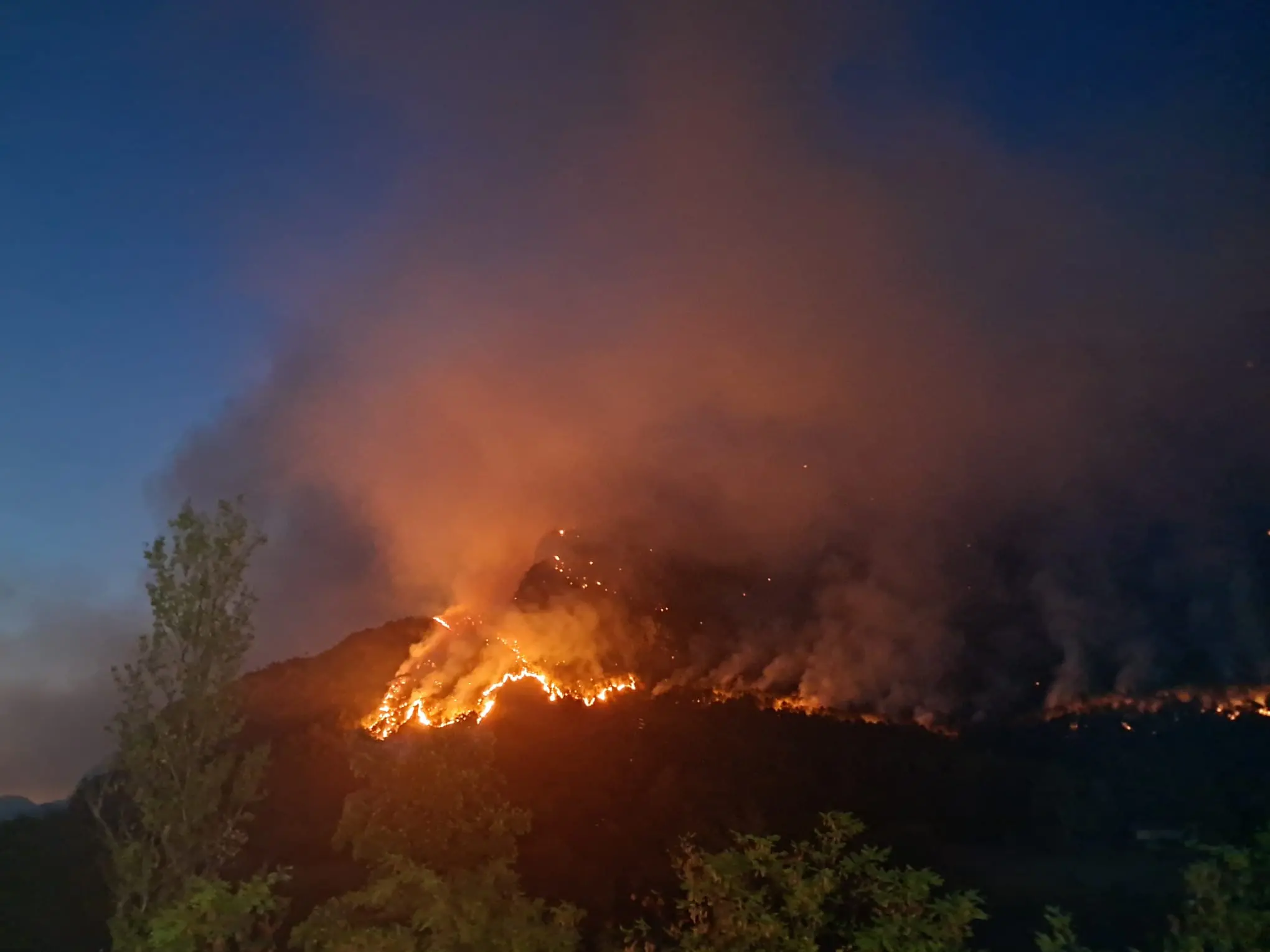 Le colonne di fumo degli incendi che stanno bruciando in Carnia sono visibili a chilometri di distanza e in alcune zone l’aria è diventata difficile da respirare. Una coltre grigia avvolge vallate e montagne, oscurando il cielo e cambiando il volto del paesaggio. Nella notte appena trascorsa, nel buio erano le fiamme sui versanti a illuminare la montagna, mentre con il giorno il fumo è tornato a dominare lo scenario.