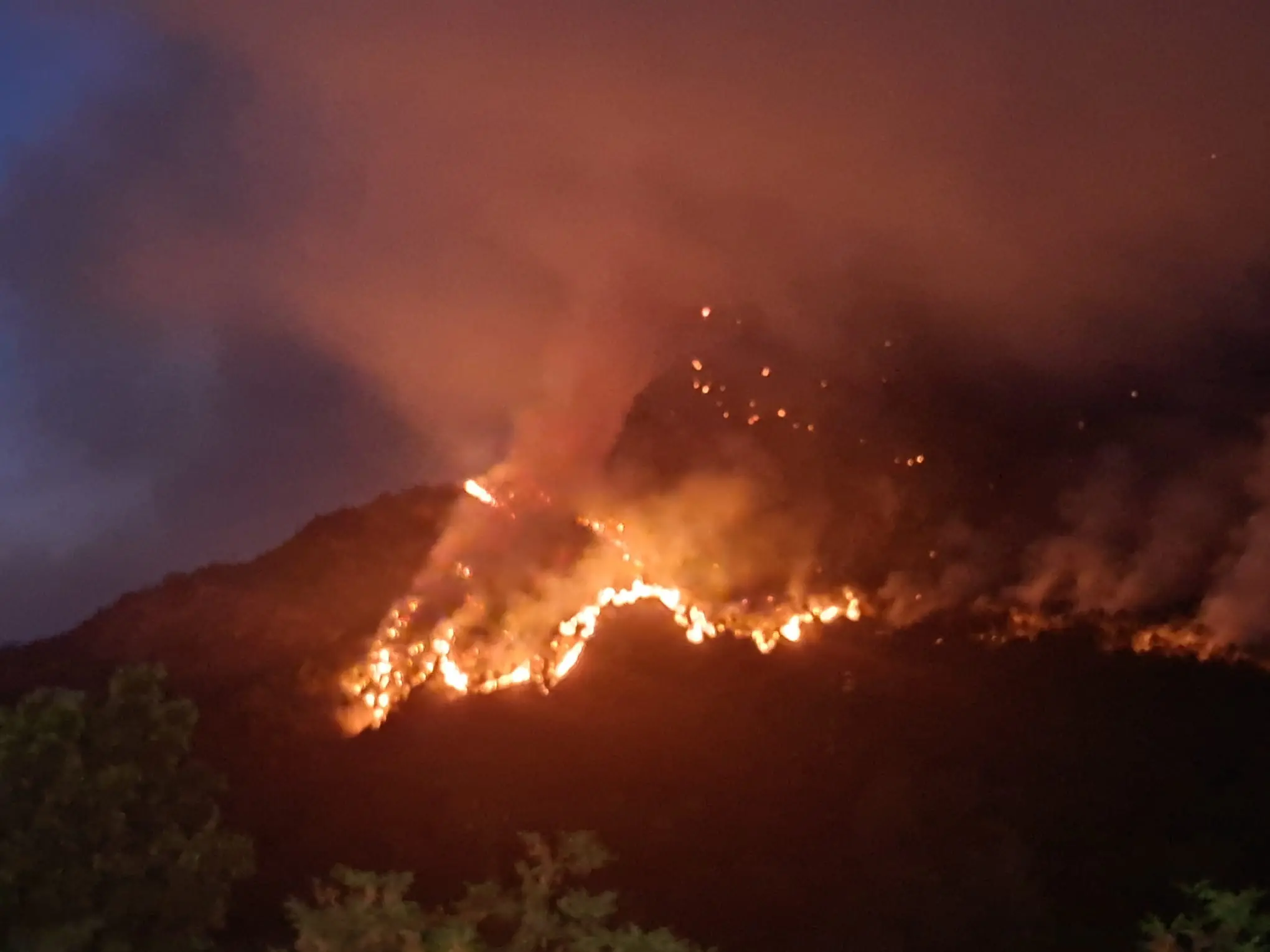 Le colonne di fumo degli incendi che stanno bruciando in Carnia sono visibili a chilometri di distanza e in alcune zone l’aria è diventata difficile da respirare. Una coltre grigia avvolge vallate e montagne, oscurando il cielo e cambiando il volto del paesaggio. Nella notte appena trascorsa, nel buio erano le fiamme sui versanti a illuminare la montagna, mentre con il giorno il fumo è tornato a dominare lo scenario.
