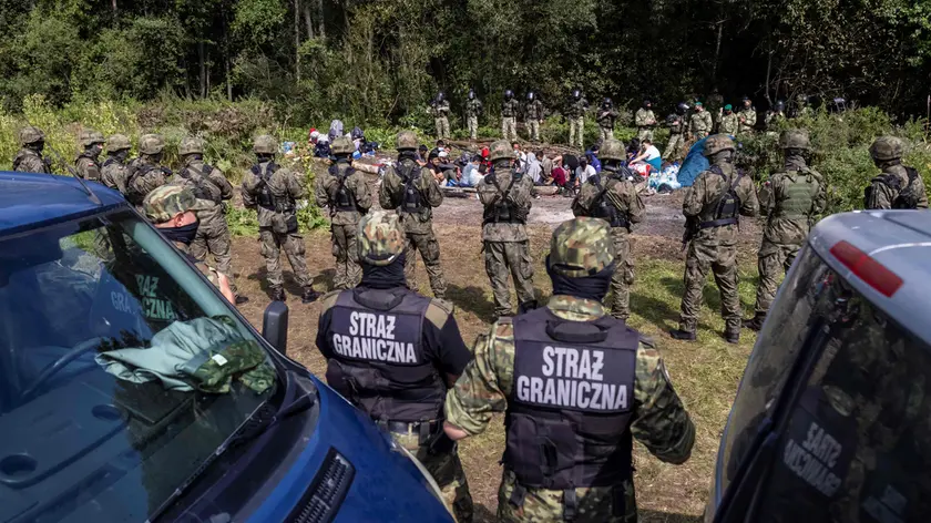 Polish border guards (foreground) stand next to migrants believed to be from Afghanistan in the small vilage of Usnarz Gorny near Bialystok, northeastern Poland, located close to the border with Belarus, on August 20, 2021. - The fate of a group of 32 bedraggled migrants stranded at a makeshift encampment on the border between Belarus and Poland for nearly two weeks has sparked a heated debate in Poland. The sight of the migrants, who are believed to be from Afghanistan, stuck between armed Belarusian officers and Polish soldiers just a few metres away has moved many Poles. (Photo by Wojtek RADWANSKI / AFP)