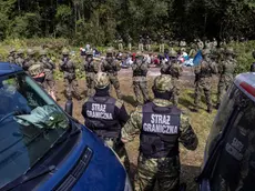 Polish border guards (foreground) stand next to migrants believed to be from Afghanistan in the small vilage of Usnarz Gorny near Bialystok, northeastern Poland, located close to the border with Belarus, on August 20, 2021. - The fate of a group of 32 bedraggled migrants stranded at a makeshift encampment on the border between Belarus and Poland for nearly two weeks has sparked a heated debate in Poland. The sight of the migrants, who are believed to be from Afghanistan, stuck between armed Belarusian officers and Polish soldiers just a few metres away has moved many Poles. (Photo by Wojtek RADWANSKI / AFP)