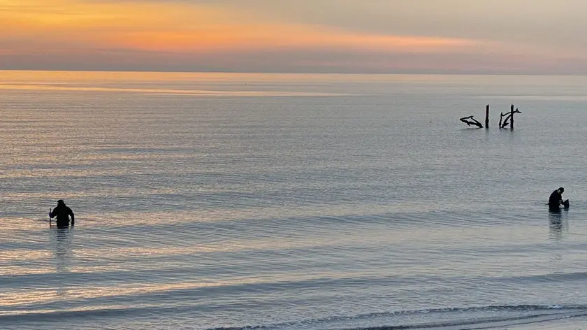 Cacciatori di oggetti muniti di metaldetector si immergono davanti alla diga al tramonto in un’immagine di Enrico Cester