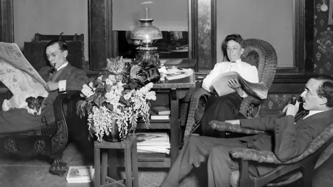 ca. 1900 --- Adults, with their cat and dog, enjoy reading and smoking in a turn of the century living room. --- Image by © Kirn Vintage Stock/Corbis