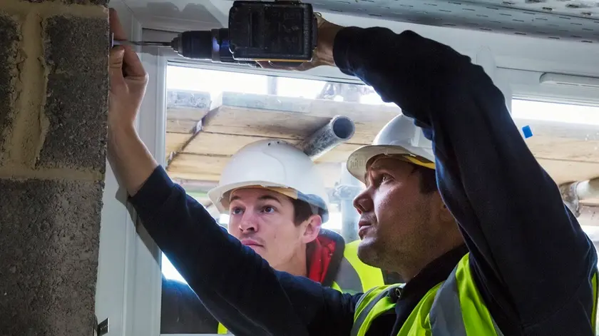 Two workmen on a construction site, builder in hard hat using an electric drill on a window frame.