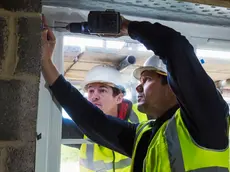 Two workmen on a construction site, builder in hard hat using an electric drill on a window frame.