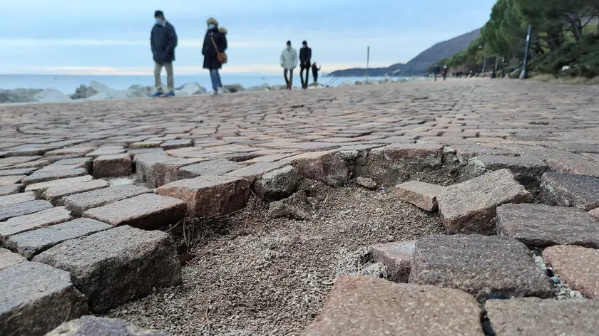 Una parte della pavimentazione in porfido danneggiata sul lungomare Benedetto Croce a Barcola e, a destra in basso, cittadini a passeggio in zona. Foto di Andrea Lasorte