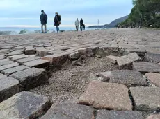 Una parte della pavimentazione in porfido danneggiata sul lungomare Benedetto Croce a Barcola e, a destra in basso, cittadini a passeggio in zona. Foto di Andrea Lasorte